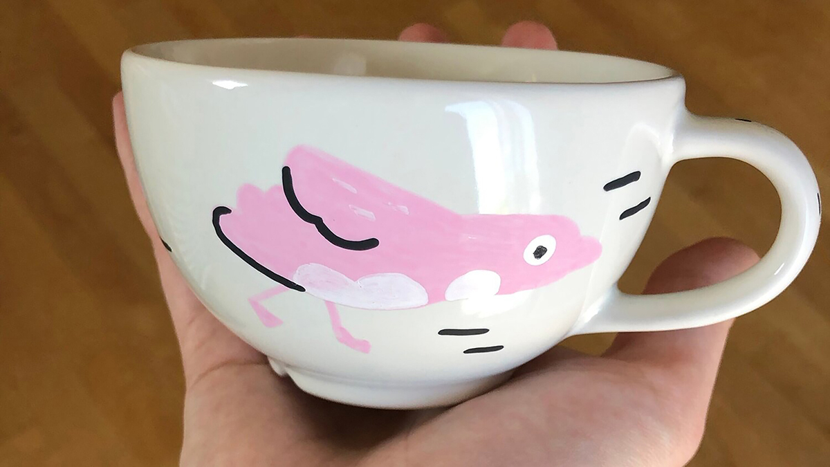 Photograph of a hand holding a ceramic mug with a pink hand painted bird on it