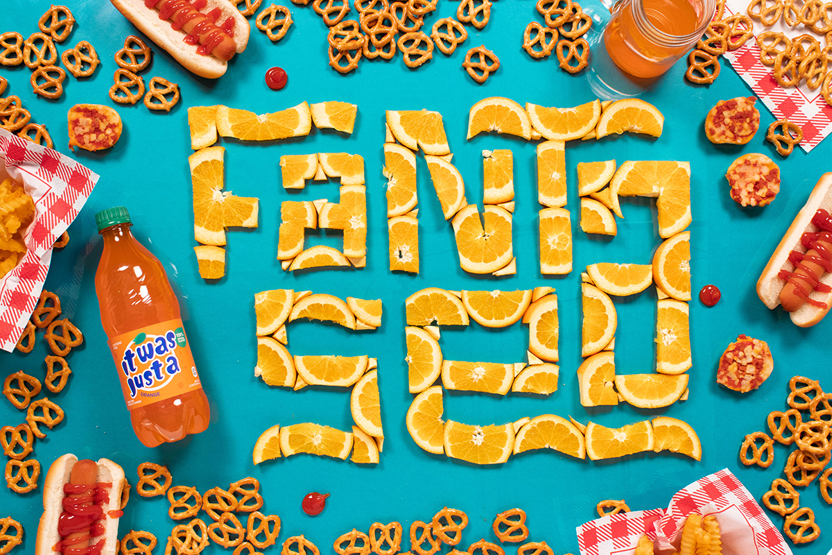 Turquoise table with red and white napkins, hot dogs, pretzels, and orange Fanta soda bottle. Oranges cut and arranged to read Fanta Sea.