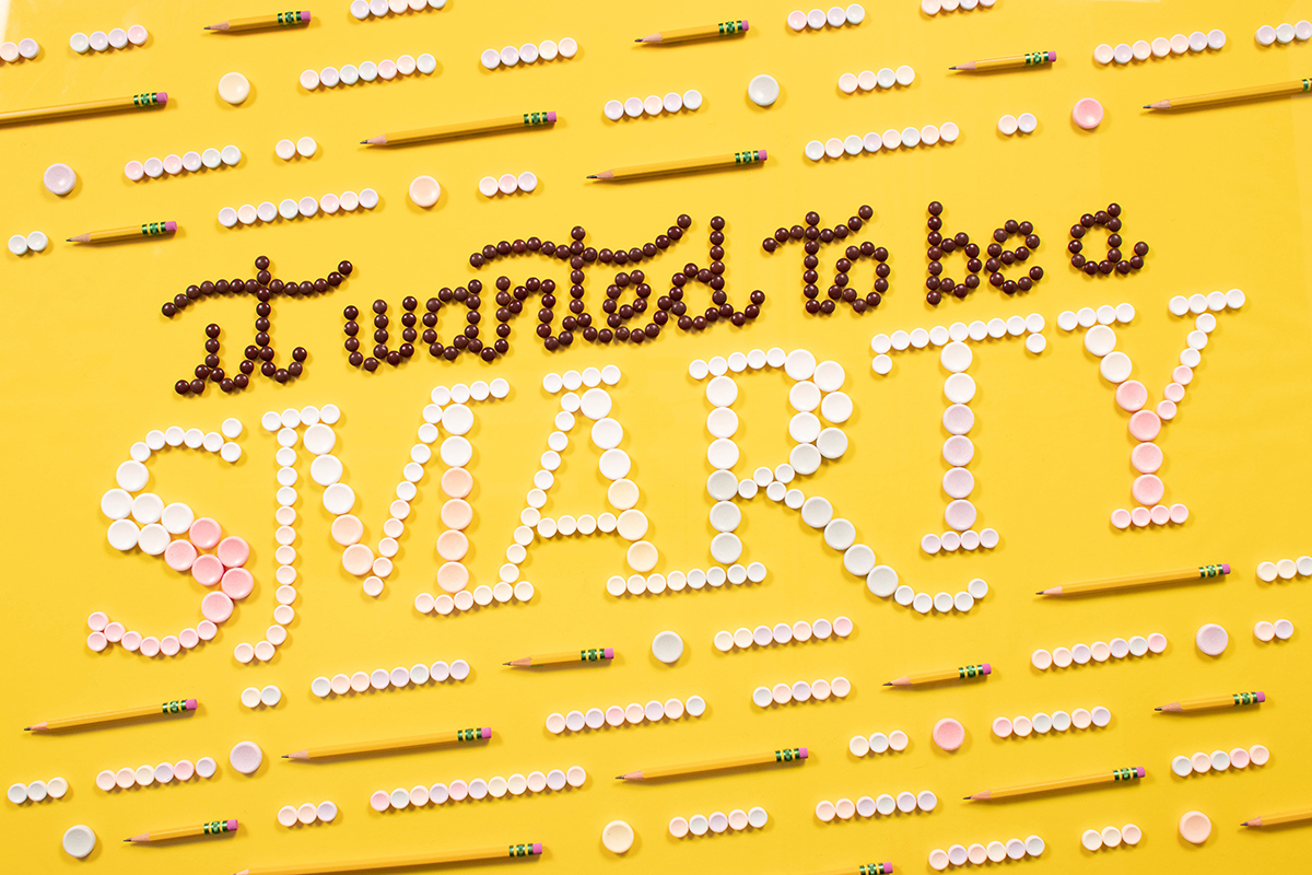 Intricate hand lettering built from Smarties candy and pencils, reads 'It Wanted to Be a SMARTY.' Pink, white, and brown colors on yellow background.
