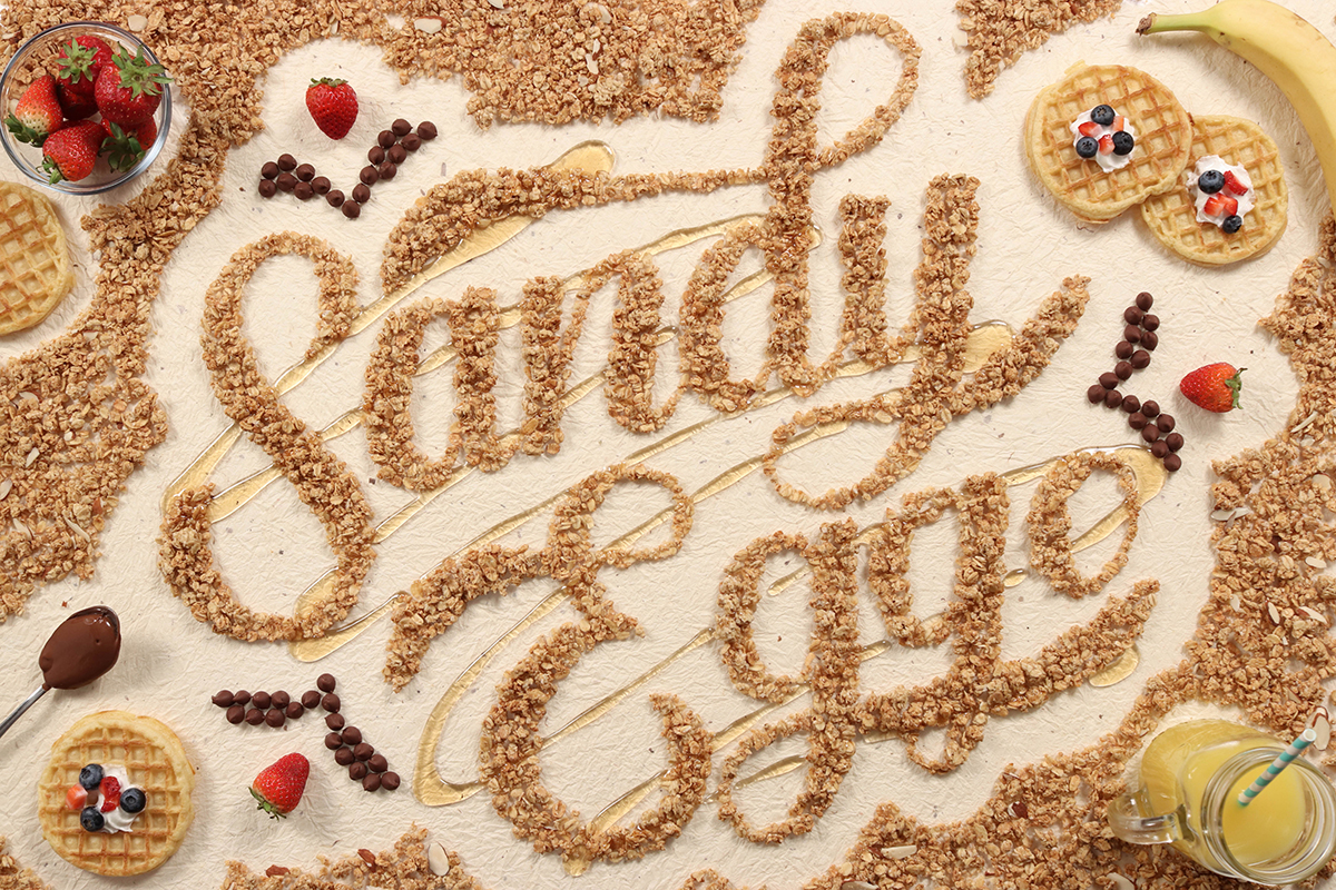Hand lettered script design shot from above, constructed of breakfast cereal, berries, honey, chocolate, and waffles. Reads 'Sandy Eggo.'