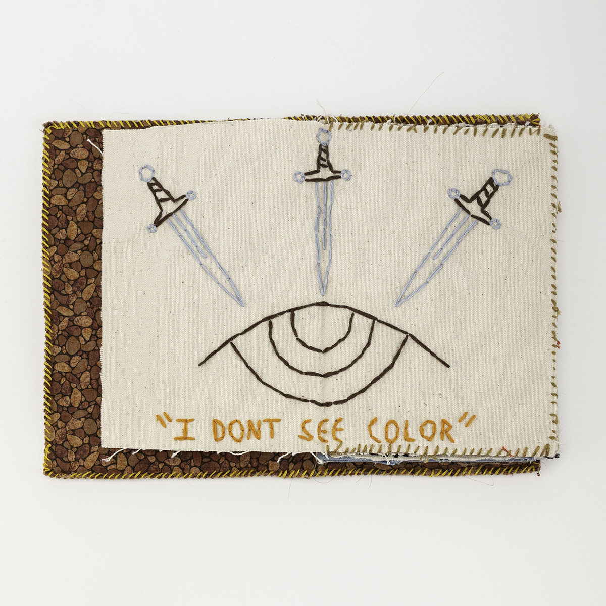Handmade book layed open two page spread with fabric pages, image of eye and three swords sewn in with sewn text below eye reading 'I don't see color'